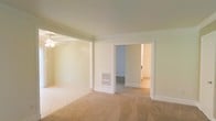 This single-floor space presents a clean, neutral palette throughout, with walls painted in soft cream tones that lend a bright, airy feel to the interior. The layout features an open connection between areas, divided by a structural column with decorative trim detail that adds subtle architectural character. One section of the space is finished with light-tiled flooring, while the adjoining area transitions to wall-to-wall beige carpet, offering a comfortable and cohesive feel across both zones. A ceiling fan is installed in the tiled area, providing both ventilation and casual lighting. A wall-mounted heating unit is positioned near the base of the dividing column, serving as a practical climate control feature. Classic white baseboards and door casings frame the openings throughout, contributing to a traditional aesthetic. A doorway leads into an additional room, visible in the background, which shares the same light tones and carpeted flooring, suggesting consistent design and finish choices throughout the space. Overall, the interior feels well-maintained, modest in scale, and move-in ready with a straightforward, functional layout.