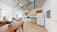 Located at 2373 E Evans Ave in Denver, CO, this 2-bedroom, 2-bathroom unit offers a distinctive industrial-modern aesthetic. Exposed concrete ceilings with visible ductwork and structural columns set a bold architectural tone throughout, contrasted by crisp white walls and light wood-style flooring that create a clean, airy atmosphere.

The open-plan living, dining, and kitchen area serves as the heart of the unit. The kitchen is equipped with light wood cabinetry, a full suite of white appliances including a range, dishwasher, microwave, and refrigerator, and a dark laminate countertop. A compact dining area with a round wooden table sits adjacent to the kitchen. The living room benefits from oversized wood-framed windows and a glass door that invite abundant natural light and offer views of the surrounding neighborhood.

Both bedrooms are bright and simply designed, each featuring exposed concrete ceilings, light flooring, and windows with outdoor views. The two bathrooms include one with a vanity featuring wood cabinetry and a wide mirror, and another with a full soaking tub and shower combination. A dedicated utility room completes the layout. The unit's industrial character, paired with its light-filled interiors, offers a functional and distinctive living environment in Denver's University Hills neighborhood.