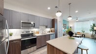 This single-floor residence offers a thoughtfully designed layout with contemporary finishes throughout. The kitchen is a standout feature, showcasing two-tone cabinetry with gray upper cabinets and rich walnut-toned lower cabinets, complemented by a marble-inspired tile backsplash and quartz countertops. A central island with bar seating provides additional prep space and casual dining, while a full suite of stainless steel appliances adds a polished, functional edge. Light wood-look flooring ties the kitchen seamlessly to the adjoining dining area.

The living room makes a bold statement with a teal textured accent wall, ceiling fan, and natural light from windows dressed with blinds and sheer curtains. The bedroom continues the design-forward approach with a vibrant botanical-print feature wall, plush carpeting, and a walk-in closet equipped with wire shelving and ample hanging space.

The bathroom is finished with large-format marble-style tile flooring, a tub-shower combination with coordinating tile surround, and a vanity with storage cabinetry. Throughout the residence, clean white walls and recessed lighting create a bright, cohesive ambiance that balances modern style with everyday comfort.