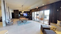 This single-floor amenity lounge in Flower Mound is designed with a contemporary aesthetic that blends dark reclaimed wood wall paneling with light large-format tile flooring throughout. The space is organized into several distinct functional zones that flow seamlessly into one another. A centerpiece billiards table anchors the main gathering area, positioned atop a blue-gray area rug and illuminated by a cluster of pendant lights suspended from the ceiling above.

Adjacent to the billiards area is a wet bar and entertainment wall clad in dark wood, featuring open shelving, a wall-mounted flat-screen television, and a pass-through window offering a view into a connected workspace equipped with computers. A built-in bench seat lined with accent pillows is positioned beneath the pass-through, accompanied by upholstered ottomans and side tables that create an informal media lounge.

Tall columns draped with sheer floor-to-ceiling curtains define a lounge corridor, adding visual softness alongside modern seating arrangements. Large glass sliding doors open directly onto an outdoor patio furnished with wooden chairs and tables, introducing abundant natural light and an easy indoor-outdoor connection. Recessed lighting throughout maintains a clean, well-lit ambiance across the entire space.