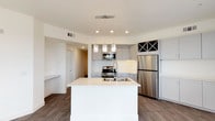 This single-floor space showcases a thoughtfully designed, open-concept kitchen that serves as the centerpiece of the layout. The kitchen is anchored by a spacious white waterfall island that provides ample workspace and doubles as a gathering point. Overhead, a trio of decorative crystal pendant lights add a touch of elegance while illuminating the island below, complemented by recessed ceiling lighting throughout.

The cabinetry is finished in a contemporary light grey tone, offering generous storage across both upper and lower units. A distinctive X-pattern wine rack built into the upper cabinetry adds a functional yet stylish detail. Stainless steel appliances, including a French door refrigerator, gas range, and built-in microwave, lend a sleek, professional quality to the space.

The island features an undermount double sink with a modern spring-neck faucet, adding practicality to its clean aesthetic. Rich, dark hardwood-style flooring runs throughout, providing a warm contrast to the bright white and grey palette. A hallway visible in the background suggests additional rooms beyond, rounding out this well-appointed, modern living space.