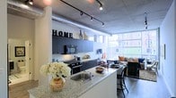 This stylish urban loft showcases a thoughtfully designed open-concept layout that seamlessly blends industrial character with modern living. Exposed concrete ceilings, visible ductwork, and track lighting run throughout the space, lending an authentic loft aesthetic that feels both functional and sophisticated.

The kitchen is a standout feature, outfitted with dark cabinetry, stainless steel appliances, and a generous granite-topped island with bar seating — ideal for both cooking and casual dining. The island's sink and ample counter space make it as practical as it is visually striking.

The open floor plan flows naturally from the kitchen into the living area, where large floor-to-ceiling windows flood the space with natural light and provide views of the surrounding urban landscape. The living area offers a comfortable and inviting atmosphere, complemented by the warmth of wood-style flooring that contrasts beautifully with the raw industrial ceiling above.

Track lighting throughout ensures the space is well-lit and adaptable, while the cohesive dark-toned finishes in the kitchen create a bold, contemporary look. Overall, this loft delivers a compelling combination of urban edge and everyday comfort, making it a distinctive and highly livable space.