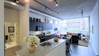 This stylish urban loft showcases a thoughtfully designed open-concept layout that seamlessly blends industrial character with modern living. Exposed concrete ceilings, visible ductwork, and track lighting run throughout the space, lending an authentic loft aesthetic that feels both functional and sophisticated.

The kitchen is a standout feature, outfitted with dark cabinetry, stainless steel appliances, and a generous granite-topped island with bar seating — ideal for both cooking and casual dining. The island's sink and ample counter space make it as practical as it is visually striking.

The open floor plan flows naturally from the kitchen into the living area, where large floor-to-ceiling windows flood the space with natural light and provide views of the surrounding urban landscape. The living area offers a comfortable and inviting atmosphere, complemented by the warmth of wood-style flooring that contrasts beautifully with the raw industrial ceiling above.

Track lighting throughout ensures the space is well-lit and adaptable, while the cohesive dark-toned finishes in the kitchen create a bold, contemporary look. Overall, this loft delivers a compelling combination of urban edge and everyday comfort, making it a distinctive and highly livable space.