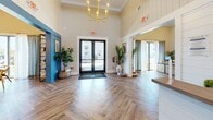 Located at 723 W 19th St in Panama City, Florida, this single-floor community space serves as a vibrant and thoughtfully designed clubhouse. The entrance greets visitors with soaring ceilings, shiplap-paneled walls, exposed dark wood beams, and an elegant gold chandelier, all set above striking herringbone-patterned hardwood flooring that flows throughout the entire space.

The living area is a standout, anchored by a bold blue diagonal-planked accent wall mounting a flat-screen TV, flanked by classic table lamps atop a neutral sideboard. Large windows dressed in sheer patterned curtains flood the room with natural light and offer outdoor views, while a glass-paneled sliding partition connects to an adjacent seating area with chess table and lounge chairs — ideal for both relaxation and social engagement.

The kitchen is equally impressive, featuring an extended run of white shaker cabinetry with matte black hardware, a generous countertop, and a dramatic herringbone blue tile backsplash. Open wood shelving displays décor and sundry items, and the space is equipped with a coffee station and small appliances, making it a fully functional amenity hub. A welcoming reception desk near the entry, branded "Welcome to the Grand Life," completes this polished and community-focused space.
