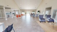 Located at 1000 SE Falcon Ln in Bentonville, Arkansas, this well-appointed single-floor facility spans a generous footprint and is thoughtfully designed for both functional and social use. The centerpiece of the space is a grand open-plan great room distinguished by soaring vaulted ceilings that create an airy, expansive atmosphere. This area features a fully equipped kitchen with gray shaker cabinetry, a teal mosaic tile backsplash, and stainless steel appliances, complemented by multiple dining tables with vibrant red chairs arranged on decorative rugs over large-format tile flooring. The living room offers a relaxed seating arrangement of upholstered wingback and accent chairs around a glass coffee table, with a wall-mounted TV, recessed lighting, and large windows that invite abundant natural light and outdoor views. The office area provides dedicated workstations with granite-topped desks, red accent chairs, and patterned carpet tile flooring, accessible through wide double doors. A separate dining room accommodates a long conference-style table surrounded by mixed seating, with windows overlooking the exterior. Throughout, the warm neutral palette, white trim, decorative artwork, and abundant natural light contribute to a bright, professional, and welcoming ambiance.