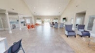 Located at 1000 SE Falcon Ln in Bentonville, Arkansas, this well-appointed single-floor facility spans a generous footprint and is thoughtfully designed for both functional and social use. The centerpiece of the space is a grand open-plan great room distinguished by soaring vaulted ceilings that create an airy, expansive atmosphere. This area features a fully equipped kitchen with gray shaker cabinetry, a teal mosaic tile backsplash, and stainless steel appliances, complemented by multiple dining tables with vibrant red chairs arranged on decorative rugs over large-format tile flooring. The living room offers a relaxed seating arrangement of upholstered wingback and accent chairs around a glass coffee table, with a wall-mounted TV, recessed lighting, and large windows that invite abundant natural light and outdoor views. The office area provides dedicated workstations with granite-topped desks, red accent chairs, and patterned carpet tile flooring, accessible through wide double doors. A separate dining room accommodates a long conference-style table surrounded by mixed seating, with windows overlooking the exterior. Throughout, the warm neutral palette, white trim, decorative artwork, and abundant natural light contribute to a bright, professional, and welcoming ambiance.