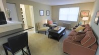 This single-floor space is thoughtfully designed with a warm, neutral color palette that creates a welcoming atmosphere throughout. The living area serves as the heart of the space, featuring plush carpeted flooring in a soft beige tone that adds comfort underfoot. A large window allows natural light to filter into the room, brightening the space and highlighting the clean, simple wall finishes. A classic radiator beneath the window underscores the space's traditional architectural character.

The living area is open and versatile, comfortably accommodating a seating arrangement that includes a sofa and a pair of accent chairs centered around a dark wood coffee table. Framed artwork on the walls adds a personal touch without overwhelming the understated aesthetic.

An open archway connects the living room to an adjacent area featuring built-in shelving, offering additional storage and display options. A breakfast bar with bar seating is also visible, suggesting a dining or kitchen area nearby. The overall ambiance is calm and functional, with classic design elements and a neutral backdrop that provides a flexible foundation for a variety of interior styles.
