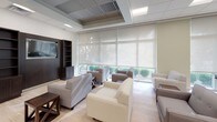 This single-floor space is thoughtfully designed to accommodate both dining and leisure, offering a clean, modern aesthetic throughout. The layout is divided into two distinct functional zones that work harmoniously together.

The dining area features multiple square tables paired with dark wood chairs with upholstered seats, arranged to comfortably seat numerous guests at once. Light wood-tone flooring runs throughout, complementing the neutral wall tones and drop-panel ceiling fitted with recessed lighting. Large windows dressed with roller shades line the perimeter, allowing generous natural light to filter in while maintaining a soft, diffused ambiance and offering views of surrounding greenery.

The lounge area provides a relaxed and welcoming atmosphere, furnished with a generous arrangement of neutral-toned sofas and armchairs grouped around low coffee tables. Floor-to-ceiling windows with sheer roller blinds flood this zone with daylight, framing outdoor views of manicured landscaping. A dark wood media unit with open shelving and a wall-mounted flat-screen television anchors one end of the lounge, adding a practical entertainment element.

A kitchenette or service area is visible at the rear of the space, along with a set of glass double doors that connect to adjacent areas. The overall design is clean, functional, and inviting, suited for community gatherings, dining, and quiet relaxation.
