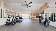 Located at 801 E Alosta Ave in Azusa, California, this fitness center offers a thoughtfully designed exercise space spread across a single, generously sized floor. The layout is divided into two distinct zones: a strength training area finished with warm wood-look flooring, and a cardio zone with durable rubber flooring, providing both aesthetic appeal and functional safety underfoot.

The strength training section is lined with floor-to-ceiling mirrored walls that visually expand the space and allow users to monitor their form. It features a multi-functional cable machine and a selection of free weights and benches, catering to a wide range of resistance training needs.

The cardio zone is bathed in natural light from large windows that frame outdoor views, creating an airy and energizing atmosphere. Treadmills, an elliptical, stationary bikes, and a rowing machine are positioned throughout this area. An oversized ceiling fan overhead ensures comfortable airflow during workouts.

A standout feature is the vibrant, large-scale abstract mural in bold orange, green, and cream tones that adorns a prominent wall, adding an artistic and motivating energy to the environment. Recessed lighting throughout ensures consistent illumination, while clerestory interior windows offer a visual connection to adjacent spaces. The overall ambiance is modern, clean, and welcoming.