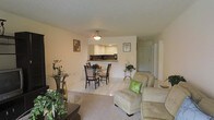 This single-floor space is designed with an open-concept layout that connects the living, dining, and kitchen areas into one cohesive, flowing environment. The interior is finished in warm, neutral tones throughout, with light-colored walls that enhance the sense of openness and brightness.

The living area is carpeted and furnished with comfortable seating, including upholstered armchairs accented with decorative throw pillows. Decorative greenery and accent pieces add warmth and character to the space. A small glass-topped side table provides a refined decorative touch near the seating area.

The dining area transitions naturally from the living space, featuring a glass-top dining table surrounded by dark-framed chairs, set atop light-toned tile flooring. A pendant light fixture overhead provides focused illumination over the dining area.

Directly beyond the dining space, the kitchen is accessible via a pass-through counter, offering a practical and visually connected layout. The kitchen features light cabinetry and dark countertops, with recessed lighting providing functional illumination.

A hallway visible in the background leads toward additional rooms, suggesting a well-organized and efficiently arranged floor plan throughout.