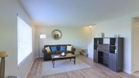 This two-story home offers a well-organized and comfortable layout designed for everyday living. The main level centers around a welcoming living room finished with light hardwood flooring and soft, neutral-toned walls that create a calm and airy atmosphere. Natural light filters in through a window, brightening the space and highlighting the clean lines of the room. A staircase with a simple railing connects the main floor to the upper level, positioned alongside a dark-toned shelving unit that adds both storage and visual contrast to the space. The living area is thoughtfully arranged, providing ample room for relaxation and casual gatherings. The overall aesthetic throughout the main level is understated and functional, with smooth ceilings and consistent flooring that ties the spaces together cohesively. The upper floor extends the living space further, offering additional rooms accessible via the interior staircase. Throughout the home, the design prioritizes a clean, livable environment with a neutral palette that serves as a versatile backdrop, making it easy to personalize. This two-floor residence balances practicality and comfort in a straightforward, well-maintained setting.