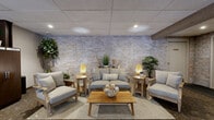 This thoughtfully designed residential lobby serves as the central hub for building residents, combining practical amenities with an inviting atmosphere. The space is anchored by a cozy lounge area featuring a light-toned stone accent wall that adds warmth and texture to the interior. Comfortable seating — including a loveseat and two armchairs in neutral upholstery with natural wood frames — is arranged around a wooden coffee table atop a layered area rug, creating an approachable gathering spot for residents. Ambient table lamps and recessed ceiling lighting provide a welcoming glow throughout. A suspended drop ceiling with a grid tile system runs throughout the space, and patterned carpet flooring adds a grounded, practical finish. Adjacent to the lounge, a bank of dark wood cabinetry provides organized, floor-labeled package storage, offering residents a secure and convenient parcel pickup solution. A glass-partitioned entryway with full-height glazing and a dark metal frame connects the lobby to the building's main entrance, where a mailbox bank and water fountain are visible. Corridor access to a stairwell and leasing office is clearly marked, with greenery accents placed throughout to soften the functional elements and enhance the overall ambiance.