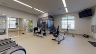 The fitness center at 800 Central in Salem, New Hampshire is a well-appointed, single-level facility designed to support a comprehensive workout experience. The space is divided into two distinct areas: a main gym floor and an adjoining movement studio, connected by a glass-paneled door that maintains an open, airy feel throughout.

The main gym area features light-toned rubber flooring and is illuminated by long linear LED light fixtures set into a clean white ceiling, providing bright, even lighting. Large windows along the walls invite natural light and offer views to the exterior. The room is outfitted with multiple treadmills, elliptical machines, a stationary bike, and a multi-function cable and weight machine, alongside a dedicated free weight section with a dumbbell rack. A striking black-and-white architectural mural serves as a bold focal point on one wall, adding character to the space.

The adjacent movement studio features warm hardwood-style flooring and soft teal accent walls, creating a calming atmosphere ideal for stretching, yoga, or group exercise. A wall-mounted flat-screen TV, full-length mirrors, and a large window with natural light make this a versatile and inviting space. Equipment such as stability balls, resistance bands, medicine balls, and foam rollers is neatly organized along the walls, ensuring the studio remains functional and uncluttered.