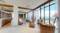 Located at 401 E Las Olas Blvd in the heart of Fort Lauderdale, this high-rise amenity floor delivers a sophisticated blend of coastal design and modern functionality. Expansive floor-to-ceiling windows wrap around the main lounge area, framing sweeping panoramic views of the Fort Lauderdale skyline and surrounding cityscape, flooding the interior with abundant natural light. Large-format porcelain tile flooring runs throughout, complemented by warm wood-clad columns and accent walls that ground the space with natural texture.

The main lounge features generously spaced wicker and upholstered seating arrangements positioned to take full advantage of the views, while a wall-mounted television and built-in cabinetry provide a media-ready nook with wood-paneled detailing. A formal dining area accommodates a large party, furnished with tufted leather chairs, dual globe chandeliers, and a bold abstract artwork as a centerpiece. Adjacent to the main gathering areas, a bar and kitchen zone offers light wood cabinetry, dark granite countertops, and stainless steel appliances. A casual lounge nook features built-in bench seating with wood tile walls, and intimate seating vignettes with botanical wall art add quiet, refined moments throughout the space.