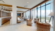 Located at 401 E Las Olas Blvd in the heart of Fort Lauderdale, this high-rise amenity floor delivers a sophisticated blend of coastal design and modern functionality. Expansive floor-to-ceiling windows wrap around the main lounge area, framing sweeping panoramic views of the Fort Lauderdale skyline and surrounding cityscape, flooding the interior with abundant natural light. Large-format porcelain tile flooring runs throughout, complemented by warm wood-clad columns and accent walls that ground the space with natural texture.

The main lounge features generously spaced wicker and upholstered seating arrangements positioned to take full advantage of the views, while a wall-mounted television and built-in cabinetry provide a media-ready nook with wood-paneled detailing. A formal dining area accommodates a large party, furnished with tufted leather chairs, dual globe chandeliers, and a bold abstract artwork as a centerpiece. Adjacent to the main gathering areas, a bar and kitchen zone offers light wood cabinetry, dark granite countertops, and stainless steel appliances. A casual lounge nook features built-in bench seating with wood tile walls, and intimate seating vignettes with botanical wall art add quiet, refined moments throughout the space.
