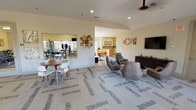 This single-floor clubhouse is designed to serve as a versatile community hub, offering a range of amenities across several distinct areas that flow seamlessly into one another. The main lounge is a generously sized gathering space anchored by patterned carpeting, comfortable modern seating including leather lounge chairs with colorful accent pillows, a wall-mounted flat-screen television, and a ceiling fan overhead. Playful typographic wall art adds a casual, welcoming personality to the room. Large windows throughout the space allow abundant natural light to fill the interior, while recessed lighting provides additional illumination. Adjacent to the lounge is an expansive game and recreation area, featuring a pool table and additional seating arrangements, all bathed in natural light from a generous bank of windows overlooking the outdoors. A mirrored fitness corner is integrated into the layout, housing exercise equipment for residents' convenience. The well-equipped kitchen is outfitted with warm natural wood cabinetry, granite countertops, a decorative tile backsplash, a center island, a sink, a coffee maker, a microwave, and under-counter refrigeration — making it ideal for hosting gatherings. Tile flooring in the kitchen transitions smoothly into the carpeted social areas, tying the clubhouse together as a cohesive, functional, and inviting community space.