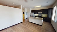 Located at Wewatta Way in Denver, Colorado, this single-floor unit offers a well-designed, open layout with modern finishes throughout. The kitchen serves as a central highlight, featuring rich dark cabinetry that extends floor to ceiling, providing generous storage capacity. A granite-topped island with an integrated sink anchors the space, complemented by stainless steel appliances including a French door refrigerator with an ice dispenser, an electric range, and a built-in microwave. Light-toned hardwood-style flooring runs throughout the main living areas, creating a warm, cohesive aesthetic that connects the kitchen to the open living space. Natural light enters through windows fitted with horizontal blinds, offering views of the surrounding area. The living area is open and versatile, with ceiling fans providing air circulation. The bedroom features plush carpeting underfoot and direct access to a spacious walk-in closet with built-in shelving and hanging rods for organized storage. The bathroom is cleanly appointed with tile flooring, a soaking tub with a tiled surround, a overhead shower, and dark wall-mounted cabinetry for additional storage. Clean white walls and neutral tones throughout lend the unit a bright, move-in-ready ambiance.