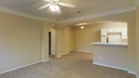 This single-story space presents an open-concept layout characterized by warm beige walls and neutral wall-to-wall carpeting that extends throughout the main living area. Crown molding frames the ceiling, adding a refined architectural detail that complements the overall design. A ceiling fan with a light fixture is centrally positioned in the main living area, providing both air circulation and ambient lighting.

A distinctive arched doorway serves as a prominent architectural feature, creating a graceful transition between the living area and an adjoining hallway or secondary room. Pilasters flank the arch, reinforcing the structured yet inviting character of the space. A secondary pendant light fixture illuminates the far end of the room, ensuring consistent lighting throughout.

To one side, a partial wall and raised countertop indicate a kitchen area, with white cabinetry and a refrigerator visible, suggesting a functional and accessible kitchen layout. The open sightlines between the kitchen and living space enhance the sense of connectivity and ease of movement. Overall, the space offers a clean, functional environment with classic design touches that maintain a welcoming and comfortable atmosphere.