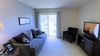 This single-floor space centers around a well-appointed living room that combines comfort with practicality. The room is finished with wall-to-wall carpet, providing a soft, inviting surface underfoot, and features a textured ceiling that adds subtle visual interest overhead. A large sliding glass door dominates one wall, fitted with vertical blinds and sheer curtains that allow natural light to filter in while maintaining privacy. The door opens to a view of a lush, green outdoor area, creating a pleasant connection between the interior and exterior. The walls are painted in a cool, muted lavender-gray tone, contributing to a calm and relaxed ambiance throughout the space. A wall-mounted piece of framed artwork adds a decorative accent. The room offers ample space for seating and entertaining, as evidenced by the arrangement of a full-sized sofa and an upholstered accent chair. A ceiling-mounted air vent ensures year-round climate comfort. Overall, the living area is bright, neutral, and functional, making it a versatile space suitable for everyday living.