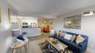 This single-story unit is thoughtfully designed with an open-concept layout that connects the living, dining, and kitchen areas into one cohesive space. The living room features wood-look plank flooring, a textured ceiling, and a large sliding glass door fitted with vertical blinds that allows natural light to filter in. A geometric-framed mirror and arc floor lamp serve as notable design accents, complementing the neutral wall tones throughout.

The kitchen is positioned behind a peninsula-style breakfast bar topped with dark countertops, providing both additional prep space and a visual separation from the living area. White cabinetry lines the walls, and stainless steel appliances—including a refrigerator, microwave, and range—equip the space efficiently. A pass-through opening connects the kitchen visually to the rest of the unit.

Adjacent to the kitchen, the dining area accommodates a round glass table and is accented by a ceiling fan and decorative wall art. The bedroom is carpeted and features a window with blinds and curtains, offering a quiet retreat. A full bathroom includes a tub-shower combination with white surrounds. A closet with shelving provides practical storage near the entryway.