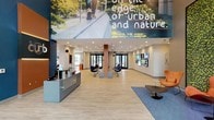 The lobby at 150 Glover Ave in Norwalk, CT makes a bold first impression with its open, airy layout and contemporary design. Upon entry, visitors are greeted by a sleek reception desk set against a striking large-scale mural that reads "on the edge of urban and nature," instantly communicating the property's identity. Light wood-style flooring runs throughout, complementing the neutral white and gray walls accented by deep navy blue feature walls.

The main lounge area is generously proportioned, furnished with an eclectic mix of mid-century modern seating, including sling-style chairs, modular sofas, and unique accent pieces such as a cowhide butterfly chair and a sculptural wood coffee table. Large windows along the perimeter flood the space with natural light and offer views of the surrounding landscape.

A vibrant communal kitchen sits within the lobby footprint, outfitted with a retro-style orange refrigerator, stainless steel appliances, marble-accented countertops, open shelving, and high-top wood dining tables — ideal for casual gatherings or quick meals.

A dedicated conference area features a rounded wood table surrounded by mustard yellow chairs beneath a bold circular pendant light, set against a dramatic navy accent wall. Display screens and a living green wall add dynamic visual interest throughout, creating an energetic yet welcoming ambiance suited for both residents and guests.