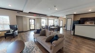 This single-story clubhouse is designed to serve as a welcoming community hub, offering a variety of functional and comfortable spaces throughout its open layout. Rich hardwood-style flooring flows consistently across the entire space, anchoring the warm, modern aesthetic.

The main gathering area features an open-concept lounge with plush seating, including a neutral-toned sofa accented with decorative pillows, arranged atop a patterned area rug. Adjacent to the lounge, a dedicated dining area offers multiple round tables paired with tufted upholstered chairs, all bathed in natural light from large windows with horizontal blinds.

A fully equipped community kitchen is outfitted with dark cabinetry, light countertops, recessed lighting, and full-size appliances including a refrigerator and microwave. The kitchen's generous counter space and pass-through bar make it ideal for hosting events or casual gatherings.

A separate lounge area provides a more intimate retreat, centered around a tile-surround fireplace topped with a wall-mounted flat-screen television. Built-in bench seating lines the walls, and navy wingback chairs flank a large interior window, creating a comfortable viewing and relaxation space.

Throughout the clubhouse, crown molding, recessed lighting, and a neutral color palette contribute to a clean, cohesive, and inviting atmosphere suited for a wide range of community activities.
