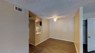 This single-floor unit presents a practical and well-organized layout designed to maximize everyday functionality. The living area is finished with rich hardwood-style flooring that extends throughout the space, adding warmth and visual continuity. A ceiling fan overhead provides both lighting and air circulation, keeping the room comfortable year-round. The textured ceiling adds a classic touch to the interior. A defining architectural feature is the pass-through window that connects the kitchen to the living area, creating an open and interactive flow between the two spaces. The kitchen, visible through the pass-through, is equipped with appliances and cabinetry, offering a compact yet efficient workspace. The walls are finished in a neutral beige tone, providing a versatile backdrop suited to a variety of furnishing styles. HVAC vents are integrated into both the walls and ceiling, ensuring consistent climate control throughout the unit. White baseboards frame the flooring neatly, adding a clean, finished look to the rooms. Overall, the space delivers a straightforward, move-in-ready environment with functional design elements that cater well to daily living needs.