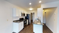 This single-floor space is designed with a clean, modern aesthetic that balances functionality and style. The kitchen serves as a standout feature, showcasing crisp white shaker-style cabinetry that extends to the ceiling for ample storage, paired with striking dark granite-style countertops that provide a bold contrast. Stainless steel appliances, including a refrigerator, microwave, and electric range, lend a contemporary and cohesive look to the cooking area. A peninsula with a built-in sink extends from the cabinetry, offering additional prep space and casual seating potential, making it ideal for both cooking and entertaining. Rich, dark hardwood-style flooring runs throughout, adding warmth and continuity to the space. Recessed and flush-mount lighting fixtures illuminate the kitchen evenly, creating a bright and inviting atmosphere. A doorway visible from the kitchen hints at additional rooms beyond, suggesting a well-connected floor plan. The overall ambiance is fresh, modern, and move-in ready, with thoughtful finishes that elevate everyday living.