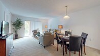 This single-floor residence offers a thoughtfully designed layout that balances comfort and functionality. The open-plan living and dining area forms the heart of the home, featuring plush wall-to-wall carpeting, neutral walls, and a textured ceiling. A pendant light fixture anchors the dining space, which accommodates a round wooden table with seating for four. The living area provides generous room for a full seating arrangement and is brightened by natural light streaming through sliding glass doors fitted with vertical blinds, offering access to an outdoor area with a glimpse of greenery beyond. The kitchen is equipped with ample white cabinetry, a tile floor, a freestanding electric range, and a full-size refrigerator, providing practical storage and workspace. The primary bedroom is spaciously sized with soft carpet, a dedicated closet, and direct access to a bathroom featuring a wide vanity, large mirror with Hollywood-style lighting, and a separate shower-tub combination. A second bathroom serves the rest of the home and includes a single-sink vanity with under-cabinet storage, dual mirrored medicine cabinets, and a full-size tub and shower. Throughout the residence, the clean, light color palette and straightforward layout create a calm and inviting atmosphere.