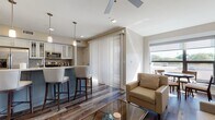 Located in the heart of Royal Oak, Michigan, this well-appointed single-floor residence offers a thoughtfully designed open-concept layout that seamlessly connects the living, dining, and kitchen areas. Rich hardwood-style flooring runs throughout the main living spaces, complemented by neutral warm-toned walls and ceiling fans that enhance the bright, airy atmosphere. Large windows and a sliding glass door with vertical blinds invite abundant natural light and provide outdoor views, giving the living area a welcoming, open feel. A built-in media wall with floating shelving anchors the living room, creating a clean, modern focal point. The kitchen is a standout feature, boasting white cabinetry with glass-front uppers, a tile backsplash, stainless steel appliances, and a curved island topped with light countertops and seating for multiple guests, illuminated by elegant pendant lighting. The bedroom is a serene retreat with plush carpeting, a ceiling fan, a spacious closet, and direct access to a well-appointed en-suite bathroom. The bathroom features large-format tile flooring, a glass-enclosed walk-in shower, and dark floating shelves for storage. The overall ambiance of this residence is contemporary and refined, offering comfort and style in a prime Royal Oak location.