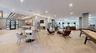 Located at 430 Englewood Ave SE in Atlanta's Grant Park neighborhood, this expansive single-floor amenity lounge offers a sophisticated and versatile communal experience. The open-plan layout is anchored by striking herringbone-patterned light wood flooring that flows seamlessly throughout, lending warmth and visual rhythm to the space. Exposed wood ceiling beams add an architectural contrast against the crisp white ceiling, while recessed lighting and sleek pendant fixtures ensure the space is well-lit throughout. Multiple seating zones are thoughtfully arranged across the floor, including curved lounge chairs, marble-top round dining tables with upholstered chairs, and a vibrant orange banquette beneath a decorative textured accent wall. A bar counter with barstool seating and a built-in wine refrigerator serves as a social focal point. A full-size billiards table anchors the entertainment area, while floor-to-ceiling windows and glass sliding doors flood the interior with natural light and offer views of an outdoor courtyard. A gallery wall showcases bold, large-format contemporary artwork, adding a creative energy to the space. An upper mezzanine level with glass railings, exposed beam ceilings, and a display shelving unit provides additional functional space and overlooks the main lounge below.