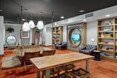 Located at 2590 Red Hill Avenue in Santa Ana, CA, this thoughtfully designed single-floor space makes a striking impression through its bold and eclectic interior aesthetic. The living room serves as the heart of the layout, showcasing rich terracotta cork flooring that adds warmth and texture throughout. A dark, moody ceiling contrasts beautifully with recessed lighting, creating an inviting and atmospheric ambiance. A standout feature is the intricate laser-cut floral accent wall, which frames two circular porthole-style window alcoves fitted with cushioned seating — offering unique, cozy nooks for relaxation. The central gathering area features a wooden dining table suspended beneath sculptural rope-hung pendant lights, complemented by woven chairs and floor cushions. A built-in bookshelf lines one wall, paired with a tufted accent chair and a wall-mounted television set within a decorative surround. The hallway connects to a well-appointed kitchenette featuring gray cabinetry, a marble-topped counter, a utility sink, and a bold botanical-patterned tile backsplash. Light wood flooring runs through the hallway, providing a clean transition between zones. Lush hanging plants add organic life to the space, completing its warm, design-forward character.