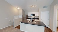 This two-floor space showcases a thoughtfully designed interior with a clean, modern aesthetic. The kitchen serves as a central highlight, featuring vaulted ceilings that lend an airy, open feel to the space. White shaker-style cabinetry lines the walls, providing ample storage, while dark countertops offer a striking visual contrast. A large central kitchen island with a double-basin sink and matching dark countertop serves as both a functional workspace and a natural gathering point. Stainless steel appliances — including a refrigerator, range with electric cooktop, and built-in microwave — complete the kitchen's contemporary look. Track lighting above the island provides focused illumination, complementing the natural light that flows through the space. The flooring transitions between hardwood-style in the kitchen and plush carpet in the adjacent areas, adding warmth and texture throughout. A staircase railing visible near the kitchen suggests a well-integrated multi-level layout, with the upper and lower floors connected seamlessly. The overall ambiance is bright, clean, and functional, making this a practical yet stylish space suited for everyday living.