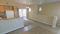 This single-floor space showcases an open-concept layout that seamlessly connects the living, dining, and kitchen areas. The living area is finished with wall-to-wall carpeting, while the dining and kitchen zones transition to hard flooring, providing a clear visual distinction between the spaces. Warm beige walls and white baseboards run throughout, creating a clean and cohesive aesthetic.

A decorative chandelier serves as a focal point above the dining area, adding a touch of elegance to the otherwise neutral interior. Natural light flows generously into the space through a large sliding glass door, which leads to an outdoor patio area and provides pleasant exterior views. A smaller arched window positioned higher on the adjacent wall adds an architectural detail while allowing additional light into the room.

The kitchen features warm honey-toned cabinetry, a granite or stone-finish countertop peninsula that partially separates it from the living area, and a white refrigerator visible along the perimeter wall. The open sightlines between all areas make the space feel expansive and functional, well-suited for everyday living and entertaining.