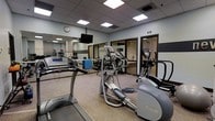 This fitness center is a dedicated single-floor facility designed to meet a variety of exercise needs. The space features a dropped ceiling with recessed panel lighting that provides bright, even illumination throughout the room. The flooring is carpeted, lending a functional and comfortable feel underfoot. A large mirrored wall on one side visually expands the space and allows users to monitor their form during workouts.

The cardio area is well-appointed with multiple treadmills and elliptical machines from recognized brands including True Fitness and Reebok, each equipped with individual digital consoles. A wall-mounted flat-screen television is positioned for easy viewing during workouts. A wall clock keeps exercisers on schedule.

Along the back of the room, a dedicated free weights and strength training area is visible, featuring a rack of dumbbells and additional equipment. Glass-paneled doors lead to an adjoining room or corridor, offering a sense of openness and connectivity within the facility.

The overall ambiance is clean, functional, and professional, reflecting a thoughtfully organized fitness environment suitable for both light and intensive training sessions.