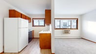 This single-floor unit is organized into a compact, practical layout that clearly delineates living, cooking, sleeping, and bathing areas. The kitchen is equipped with warm-toned wood cabinetry extending from floor to ceiling, light countertops, a stainless steel sink with a modern gooseneck faucet, and a gas range. Light hardwood-style flooring runs throughout the kitchen, and a window above the sink allows natural light to filter in. A peninsula with additional cabinetry serves as a dividing element between the kitchen and the adjoining living area.

The living space features neutral carpet flooring, white walls, and a wall-mounted air conditioning unit beneath a wide window. Baseboard heating runs along the perimeter, providing consistent warmth. The entry area transitions between carpeted and hardwood-style surfaces, with a solid entry door and flush ceiling lighting overhead.

The bedroom is carpeted and includes a built-in closet with louvered bifold doors and integrated shelving, offering practical storage. A baseboard heater lines one wall.

The bathroom is finished with dark tile flooring, white subway tile surrounding the bathtub and shower combination, a wood-front vanity with a white basin, and a mirror above. The space is clean and functional in its design.