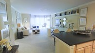 This single-floor residence showcases a thoughtfully designed open-concept layout that blends style and functionality. The expansive living area is anchored by plush, neutral-toned sofas and accented with decorative throw pillows, while a bold black-and-white checkered ottoman adds a striking visual contrast. Large floor-to-ceiling windows draped in sheer white curtains flood the space with natural light, creating an airy and inviting ambiance. Crown molding along the ceilings adds a refined architectural touch throughout. The living space flows seamlessly into a well-appointed entertainment area featuring a wall-mounted flat-screen television, open display shelving lined with decorative vases, books, and mirrors, including a standout ornate oval mirror that adds a classic elegance. A kitchen island with a dark countertop and bar seating defines the transition into the kitchen, which features light cabinetry and a clean, modern finish. The dining area sits adjacent, completing the open floor plan. The neutral color palette of soft creams and warm beiges, paired with carefully curated décor, gives the entire space a polished and sophisticated character throughout.