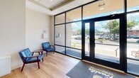 The lobby at 2405 Whitney Ave in Hamden, Connecticut serves as the welcoming entry point for Franklin Communities, offering a well-appointed and thoughtfully designed communal space. Floor-to-ceiling glass panels and double glass doors flood the entry area with abundant natural light while providing clear views of the surrounding streetscape along Whitney Avenue. Light oak hardwood-style flooring runs throughout, lending warmth and continuity to the space.

Just inside the entrance, a branded Franklin Communities mat greets visitors, with a small seating area featuring classic blue upholstered chairs positioned for comfort and convenience. The space transitions into an elevator lobby, where two elevators are flanked by a sculptural, layered ceiling fixture that adds a modern, decorative touch. A branded area rug anchors this zone.

A striking staircase with marble-effect steps, patterned blue risers, and sleek glass-panel railings with stainless steel hardware serves as a functional and architectural centerpiece. The staircase leads to an upper landing where a bank of resident mailboxes is neatly integrated into a warm wood-toned surround, accompanied by lush potted greenery that softens the space. Recessed lighting and a drop tile ceiling complete the polished, professional atmosphere throughout this compact but efficiently organized lobby.
