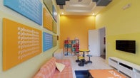 This energetic single-floor space makes an immediate impression with its bold, sunny color palette of warm yellows and vivid oranges that coat the walls from floor to ceiling. The main lounge area is a lively, well-designed gathering space anchored by a plaid orange sofa, colorful pouf seating, and a distinctive industrial-style coffee table bearing the "Esprit Loft" motif. Two flat-screen televisions are mounted side by side on a yellow accent wall above a crisp white media console, making this a functional entertainment hub as well.

A striking arched window floods the space with natural light, framing an outdoor greenery view and adding an architectural elegance that contrasts beautifully with the room's playful interior design. Horizontal orange-and-white stripes on one wall further amplify the dynamic visual energy.

A grid of colorful canvas art panels dominates another wall, each featuring motivational hashtags such as #photooftheday, #chasingyourdreams, and #nothingisimpossible, rendered in bold typography across blue, green, yellow, and orange backgrounds. A white cubby shelving unit stocked with colorful storage bins adds both practicality and visual interest. Light-toned flooring and a coffered ceiling with dark accent panels complete the space, balancing the vivid colors with clean, structured architectural details.