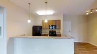 This single-floor 2-bedroom, 2-bathroom residence offers a well-designed layout that balances comfort and functionality. The open-concept kitchen serves as a central hub of the home, featuring warm honey-toned wood cabinetry that provides ample storage, granite countertops, and a full suite of black appliances including a refrigerator, electric range, and over-the-range microwave. A peninsula-style breakfast bar extends the counter space and creates a natural divide between the kitchen and the living area, making it ideal for casual dining or entertaining. Pendant lights hang above the bar, adding a stylish, layered lighting element that complements the recessed ceiling fixture overhead. The neutral color palette throughout — with soft warm walls and light ceilings — creates a bright and inviting ambiance. The layout connects seamlessly to additional living spaces, with a glimpse of the broader floor plan visible beyond the kitchen, hinting at the generous and cohesive flow of the home. The two full bathrooms and two well-proportioned bedrooms round out a practical and comfortable living environment suited for a variety of lifestyles.