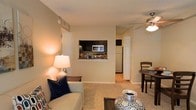This single-floor residential unit offers a thoughtfully designed open-concept layout that seamlessly connects the living, dining, and kitchen areas into one cohesive and functional space. The living area features warm, neutral-toned walls and plush carpeted flooring, creating a comfortable and inviting atmosphere. A ceiling fan with a light fixture is positioned overhead, providing both air circulation and ambient lighting to complement the soft glow of accent lamps.

A defining architectural feature of the space is the pass-through window connecting the living area to the kitchen, allowing for easy interaction between the two spaces while maintaining a sense of visual openness. The kitchen is equipped with dark cabinetry, a stainless steel microwave, and a full-size refrigerator, and features hard surface flooring that contrasts with the carpeted living and dining areas.

Adjacent to the living room, the dining area accommodates a compact round table with seating, making it well-suited for everyday dining. A tall pantry or storage closet is visible near the kitchen entry, adding practical storage to the layout. The overall ambiance of the space is warm and understated, with a neutral color palette that lends itself to a variety of décor styles.