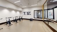 Located in Colorado Springs, CO, this professional-grade fitness facility spans a generous single floor and is divided into two well-appointed gym spaces designed to support a full range of workout disciplines. The main gym area is architecturally distinguished by soaring vaulted ceilings with exposed dark wood beams, large windows that invite abundant natural light, and sleek linear lighting throughout. This expansive zone houses a extensive row of cardio equipment — including treadmills and ellipticals — alongside a comprehensive selection of selectorized strength machines and free weights, all arranged for efficient use and easy navigation. Bold, artistic wall murals featuring dynamic geometric and nature-inspired patterns add visual energy to the space. A dedicated functional training zone includes a multi-station rig with a heavy bag, weight benches, dumbbell racks, and medicine balls, accented by large mirrors and a "Training Zone" display. A separate studio room provides a focused environment for cycling classes, equipped with spin bikes, ballet barres, floor-to-ceiling mirrors, and a wall-mounted TV. An additional alcove area accommodates rowing machines, air bikes, and supplemental free weights with its own artistic wall treatment. Throughout, the facility maintains a clean, modern aesthetic with neutral flooring, recessed lighting, and thoughtful equipment placement.
