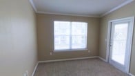 This single-story space presents a clean, well-maintained room finished with a warm, neutral color palette. The room features wall-to-wall carpeting in a speckled beige tone, providing a soft and comfortable underfoot feel. A large, multi-pane window serves as the focal point of the space, fitted with horizontal blinds that allow for adjustable natural light control. The window offers views of the surrounding exterior, bringing a sense of openness to the room. Crown molding runs along the ceiling perimeter, adding a refined architectural detail that elevates the overall aesthetic. The walls are painted in a soft tan hue, complementing the light-colored baseboards and ceiling. An electrical outlet is conveniently positioned along one wall, and the room's simple, uncluttered layout provides flexibility for a variety of furniture arrangements. The overall ambiance is calm and functional, with a modest yet polished character suited for use as a bedroom or flexible living space.