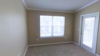 This single-story space presents a clean, well-maintained room finished with a warm, neutral color palette. The room features wall-to-wall carpeting in a speckled beige tone, providing a soft and comfortable underfoot feel. A large, multi-pane window serves as the focal point of the space, fitted with horizontal blinds that allow for adjustable natural light control. The window offers views of the surrounding exterior, bringing a sense of openness to the room. Crown molding runs along the ceiling perimeter, adding a refined architectural detail that elevates the overall aesthetic. The walls are painted in a soft tan hue, complementing the light-colored baseboards and ceiling. An electrical outlet is conveniently positioned along one wall, and the room's simple, uncluttered layout provides flexibility for a variety of furniture arrangements. The overall ambiance is calm and functional, with a modest yet polished character suited for use as a bedroom or flexible living space.