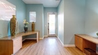 Located at 80175 Avenue 52 in La Quinta, California, this single-room office space offers a thoughtfully designed and welcoming work environment. The space is defined by a soothing soft-blue wall palette and warm light wood flooring that together create a calm, professional atmosphere.

The most striking architectural feature is a large-scale, full-wall botanical mural rendered in soft pinks, greens, and creams, serving as a vivid focal point behind the primary workstation area. Two built-in granite-topped desks with light wood cabinetry line the walls, providing functional workspace for multiple users. Additional counter space and storage cabinets continue along the perimeter, maximizing utility within the compact footprint.

Framed artwork adorns the walls alongside a gold-framed mirror, contributing to the refined aesthetic. Natural light filters in through a window fitted with a light-diffusing shade, while recessed ceiling lighting ensures consistent illumination throughout. Decorative touches — including tall ceramic vases, potted greenery, and a table lamp — add warmth and a welcoming character to the space.

A glass-panel entry door connects the office to the exterior, allowing views of the outdoors and reinforcing the bright, airy feel. Overall, this office space balances practical functionality with an inviting, artfully curated design.