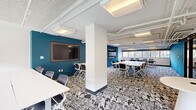 This single-floor commercial space offers a generously sized, open-plan layout designed to support collaborative work, meetings, and training sessions. The interior is organized into multiple distinct seating zones, each furnished with modern white tables and ergonomic mesh task chairs that can be reconfigured to suit a variety of group sizes and event formats.

Bold teal accent walls serve as a defining design element throughout the space, providing visual anchors for two wall-mounted flat-screen displays—ideal for presentations or video conferencing. The remaining walls are finished in clean white, reinforcing a bright, professional atmosphere. A central structural column subtly divides the floor into functional zones without fully enclosing them, preserving an open, collaborative feel.

Underfoot, a striking geometric patterned carpet in black, white, and grey adds texture and visual interest to the space. The ceiling features an exposed industrial aesthetic with white-painted structural elements, track lighting, and recessed panel fixtures that provide consistent, even illumination. A row of windows along one side of the space admits natural light and exterior views, further enhancing the openness of the environment. Wall-mounted climate control units ensure year-round comfort throughout the space.