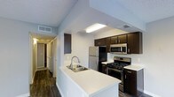 This single-story residence showcases a thoughtfully designed kitchen as its centerpiece. The kitchen features a peninsula with a built-in stainless steel sink and white countertops, providing both additional workspace and a natural division between the cooking area and the rest of the home. Dark espresso-toned cabinetry lines the walls, offering generous storage and a striking contrast against the light countertops and white walls. Stainless steel appliances, including a gas range, over-the-range microwave, and a full-size refrigerator, lend a sleek, modern aesthetic throughout. Dark hardwood-style flooring runs through the space, adding warmth and visual continuity. The textured ceiling and recessed lighting fixture above the kitchen provide functional illumination. A hallway visible from the kitchen leads further into the home, suggesting a practical, linear floor plan with additional rooms beyond. The overall design balances contemporary finishes with everyday functionality, making it a well-appointed and efficient living space.
