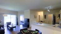 This single-floor residence offers a well-designed open-concept layout that integrates the kitchen, dining, and living areas into one cohesive, flowing space. The kitchen features a peninsula-style counter with a double sink and dark countertops, providing both functionality and a natural division between the cooking and living zones. Crown molding along the ceiling adds a refined architectural touch, while the neutral, light-toned walls create a bright and welcoming ambiance throughout. Hardwood-style flooring extends across the main living area, contributing to a warm and consistent aesthetic. The dining area accommodates a round table with seating, positioned conveniently adjacent to the kitchen for easy entertaining. The living area offers space for media and seating, with a television stand and media console visible against the central dividing wall. Natural light filters in through large vertical-blind-covered sliding doors, hinting at outdoor access. A glimpse into the bedroom reveals a well-lit, spacious retreat with ample natural light. Recessed lighting and flush-mount ceiling fixtures provide layered illumination throughout, ensuring the space feels comfortable and well-lit at all times.