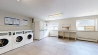 Located at 65 Paisley Blvd W in Mississauga, Ontario, this shared laundry facility offers a clean, well-organized space designed for practical, everyday use. The room is finished with large-format light-toned floor tiles throughout, contributing to a bright and hygienic atmosphere. Overhead fluorescent lighting supplements the natural light that streams in through multiple windows along the perimeter walls, keeping the space well-lit at all times.

The facility is equipped with a row of front-loading washing machines along one wall, complemented by a bank of stacked dryers, providing ample capacity for building residents. A dedicated payment kiosk is conveniently situated near the entrance for easy access. A utility sink is also available for hand-washing or pre-treatment needs.

On the opposite side of the room, a generously sized folding and sorting counter with a stone-look surface runs beneath the windows, offering practical workspace for residents. Open shelving beneath the counter provides additional storage. A bulletin board mounted on the wall displays notices and building information. Baseboard heaters ensure the space remains comfortable year-round. Seating is available with a wall-mounted bench, and the open floor plan allows for easy movement throughout the room. The overall environment is clean, functional, and well-maintained.