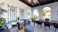 This single-story property showcases a beautifully designed covered outdoor terrace that serves as a standout architectural feature. The terrace is defined by soaring ceilings clad in rich, dark-stained wood planks, lending warmth and contrast against the bright white walls. A series of elegant arched openings lines one side of the space, framed by wrought iron balcony railings, allowing generous natural light to flood the area while offering open views of the lush tropical landscape beyond.

The terrace is thoughtfully arranged into two functional zones. A seating area features woven rattan lounge chairs paired with a tufted blue ottoman and a glass-topped side table, creating a relaxed setting for casual gatherings. Adjacent to this, a dining area accommodates a substantial wooden table surrounded by sleek black rope-woven chairs, ideal for outdoor meals.

Decorative wrought iron wall panels with intricate scrollwork add an artful, Mediterranean character to the space, complementing the overall design aesthetic. Potted tropical plants are positioned throughout, enhancing the connection to the outdoor environment. Light tile flooring ties the space together, reflecting natural light and contributing to an airy, resort-like ambiance throughout the terrace.